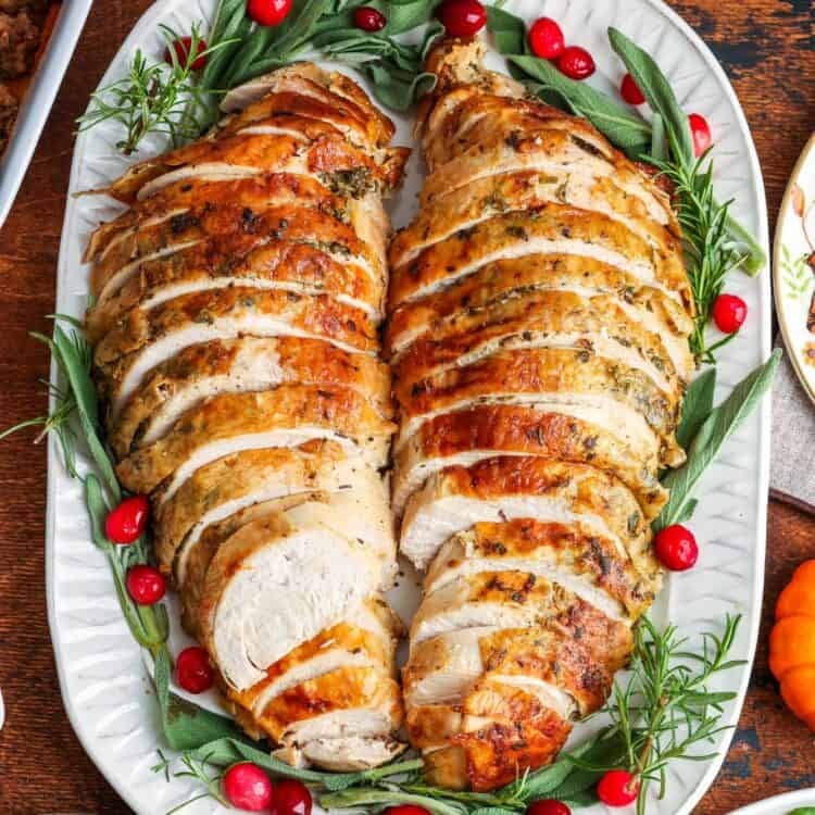sliced meat from a bone-in turkey breast on a white platter, garnished with fresh green herbs and cranberries.