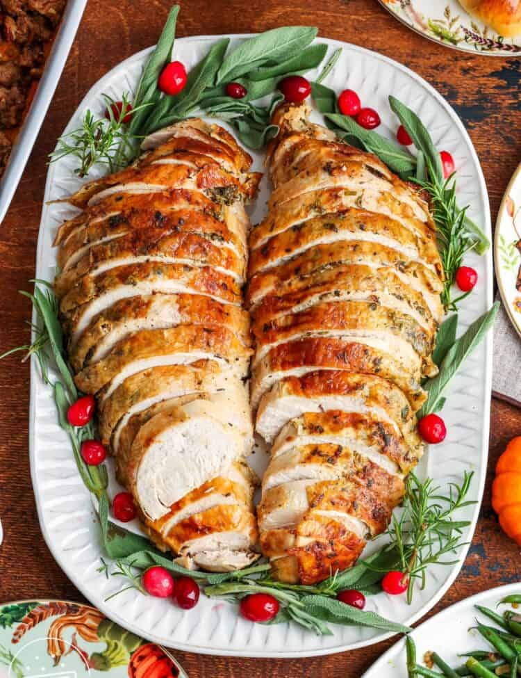 sliced meat from a bone-in turkey breast on a white platter, garnished with fresh green herbs and cranberries.