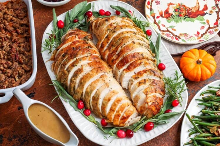 two sliced turkey breast sections on a white platter, garnished with sage leaves and cranberries. Around the turkey is a boat of gravy, a plate of green beans, and a pan of stuffing.