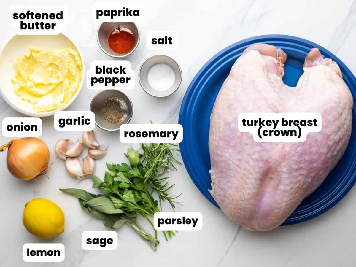 a raw turkey breast on a blue plate. Next to the turkey is softened butter, garlic, lemon, herbs, and seasonings.
