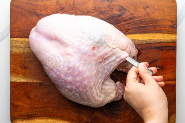 a spoon is used to gently separate the skin from the meat of a raw turkey breast that is on a wooden cutting board.