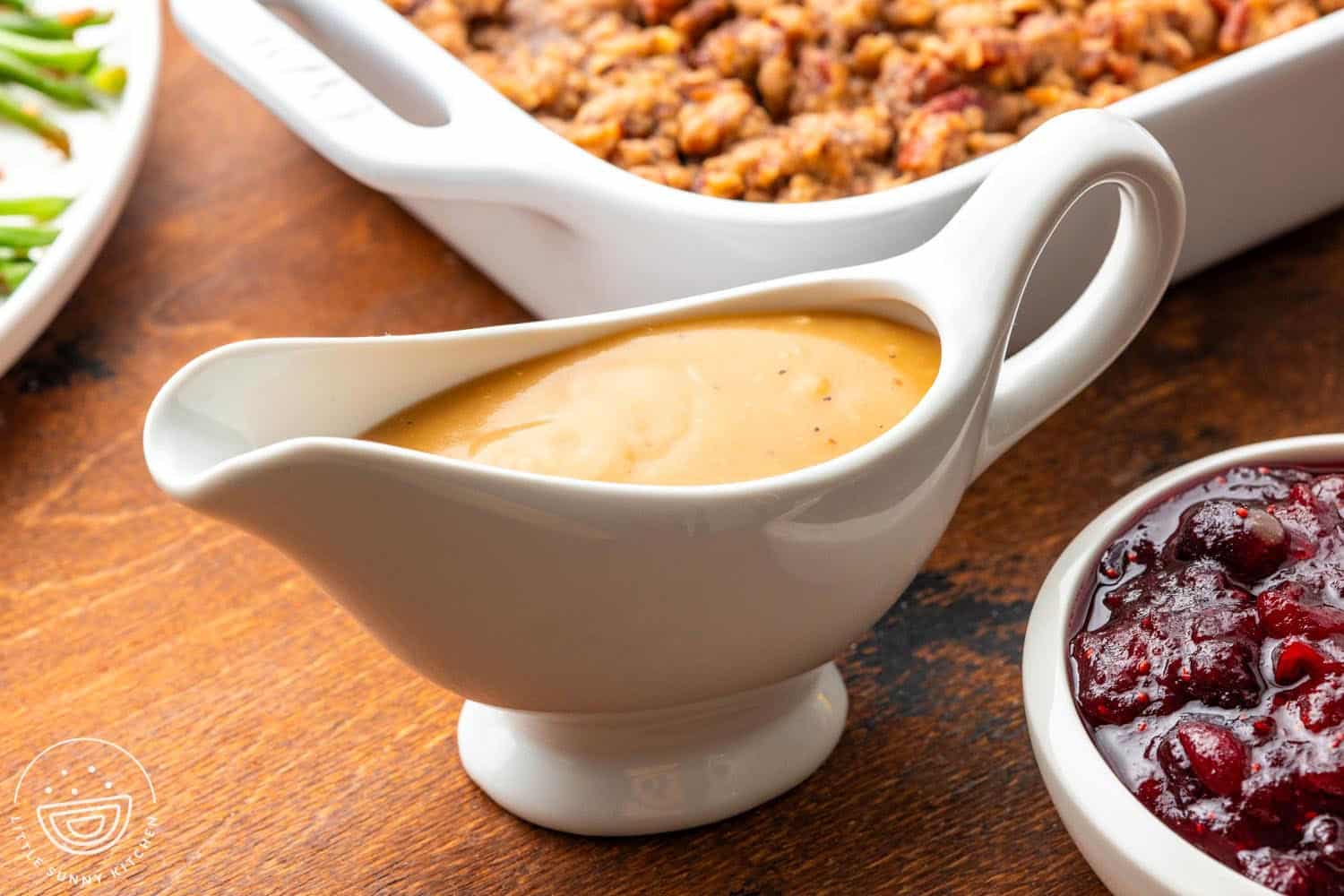a white ceramic gravy boat of homemade turkey gravy. In front of the gravy is a bowl of cranberry sauce, and behind it is a pan of stuffing.