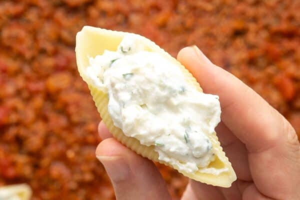 a hand holding a large pasta shell filled with creamy cheese filling.