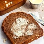 A slice of brown bread spread with butter on a sheet of parchment paper with more slices on a wooden board in the background.