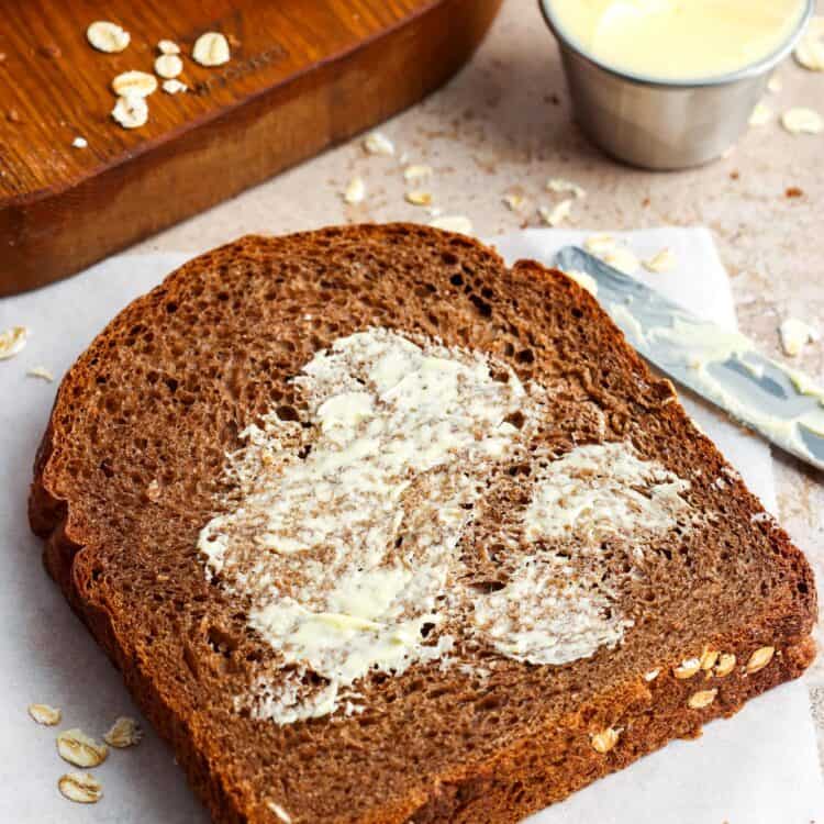 A slice of brown bread spread with butter on a sheet of parchment paper with more slices on a wooden board in the background.