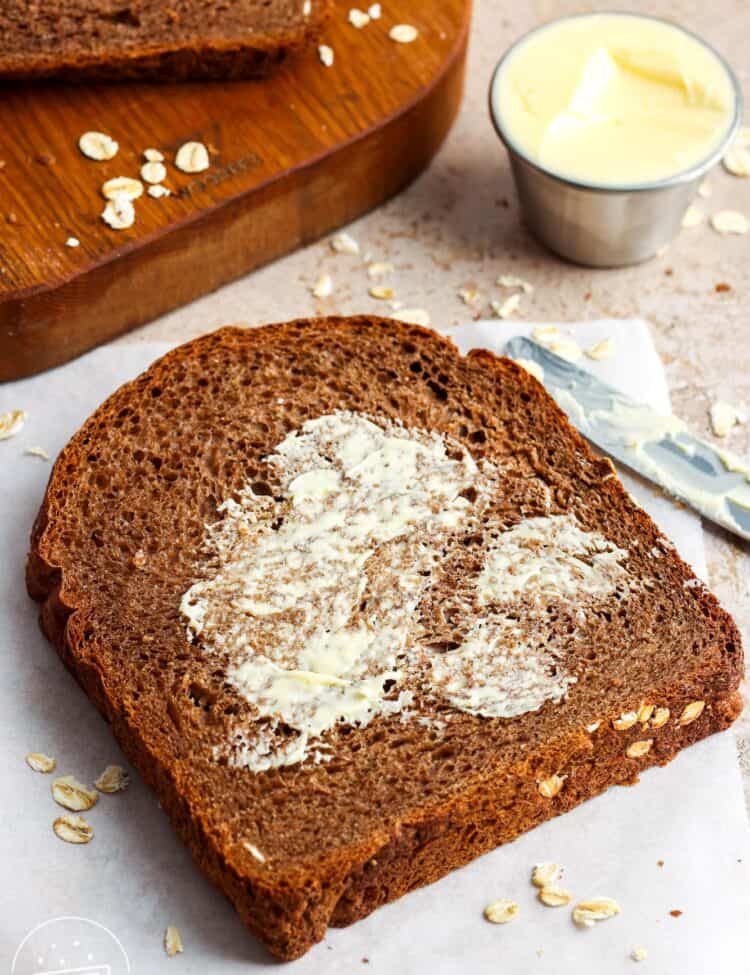 A slice of brown bread spread with butter on a sheet of parchment paper with more slices on a wooden board in the background.