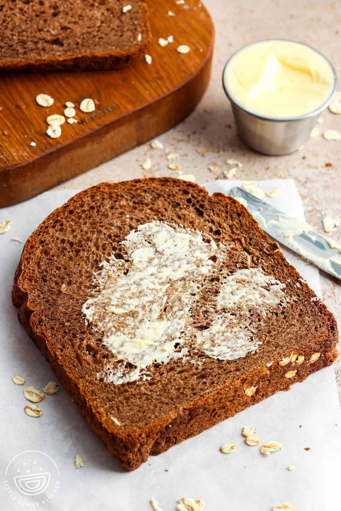 A slice of brown bread spread with butter on a sheet of parchment paper with more slices on a wooden board in the background.