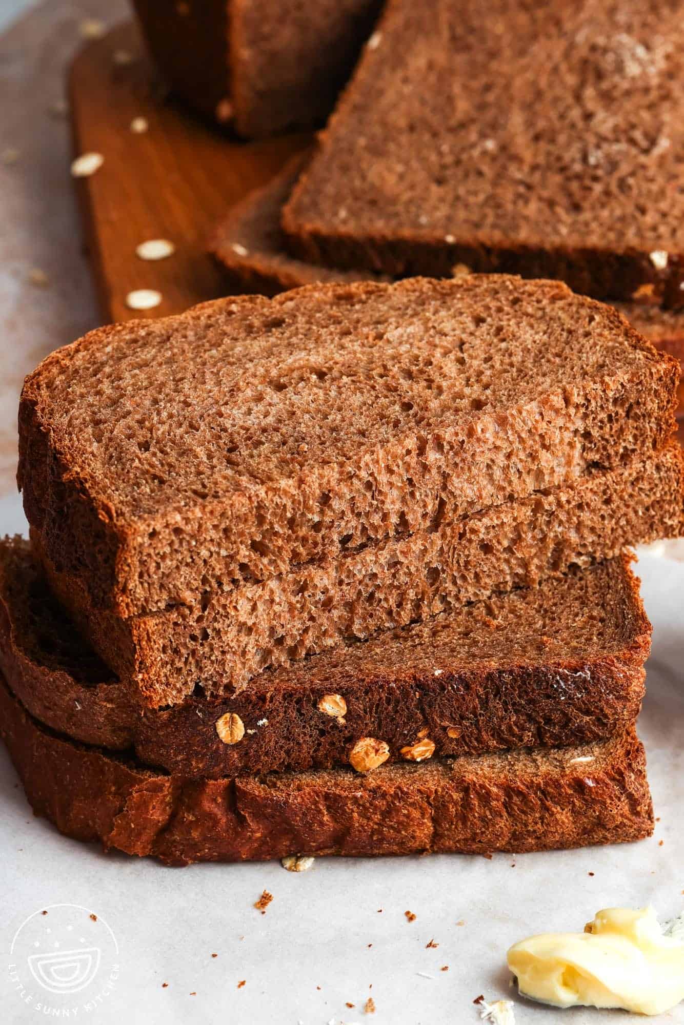 A stack of sliced brown bread showing a soft crumb and oats along the crust with a smear of butter nearby.