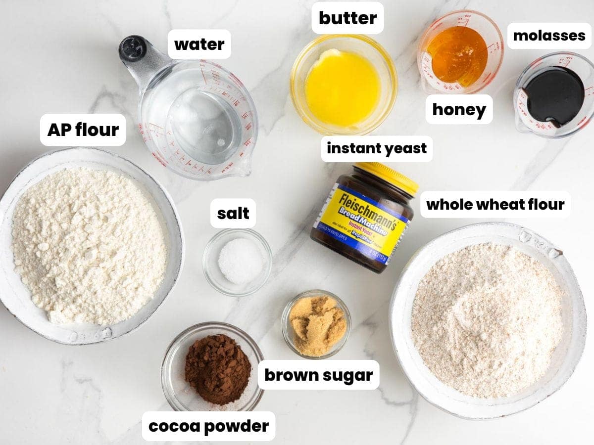 Ingredients needed to make brown bread in the bread machine including flours, water, butter, honey, molasses, brown sugar, cocoa powder, salt, and instant yeast.