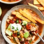 a ceramic bowl of chicken tortilla soup, topped with fried tortilla strips, sour cream, and diced avocado. There is a spoon in the bowl.