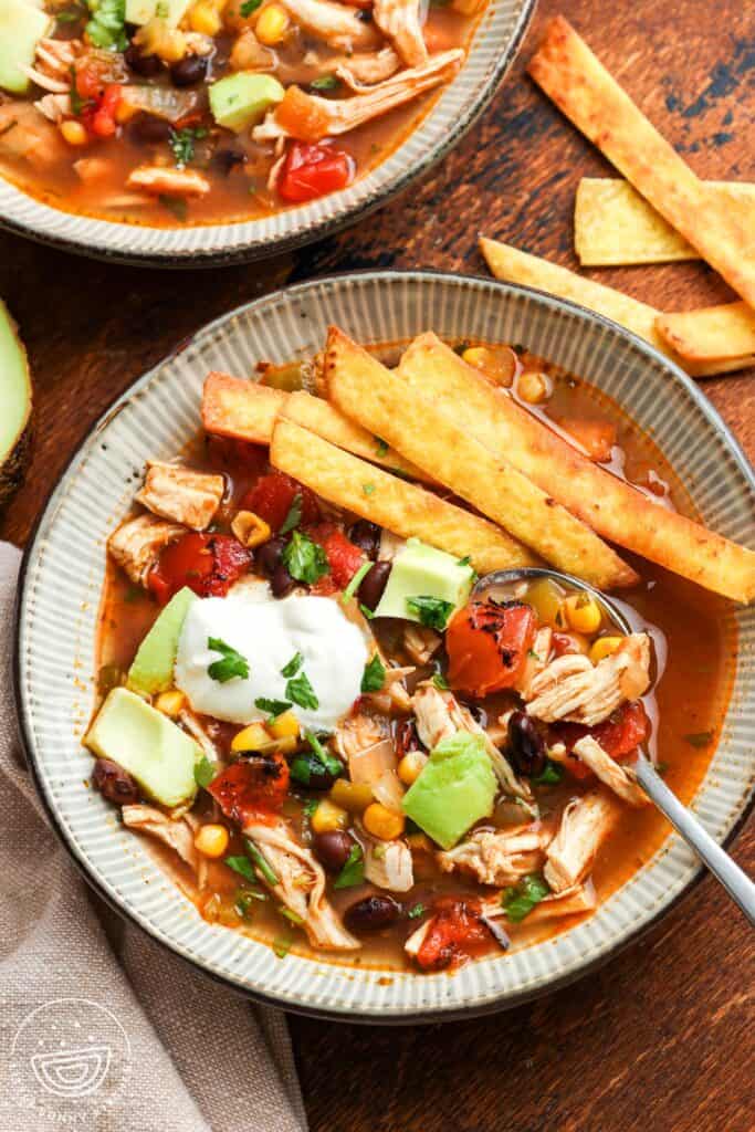a ceramic bowl of chicken tortilla soup, topped with fried tortilla strips, sour cream, and diced avocado. There is a spoon in the bowl.