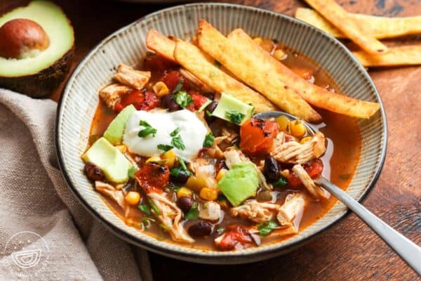 closeup of a bowl of chicken tortilla soup topped with sour cream and diced avocado, on a wooden table. In the background is half an avocado and extra fried tortilla strips.
