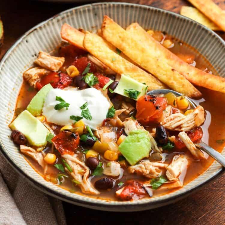 closeup of a bowl of chicken tortilla soup topped with sour cream and diced avocado, on a wooden table. In the background is half an avocado and extra fried tortilla strips.