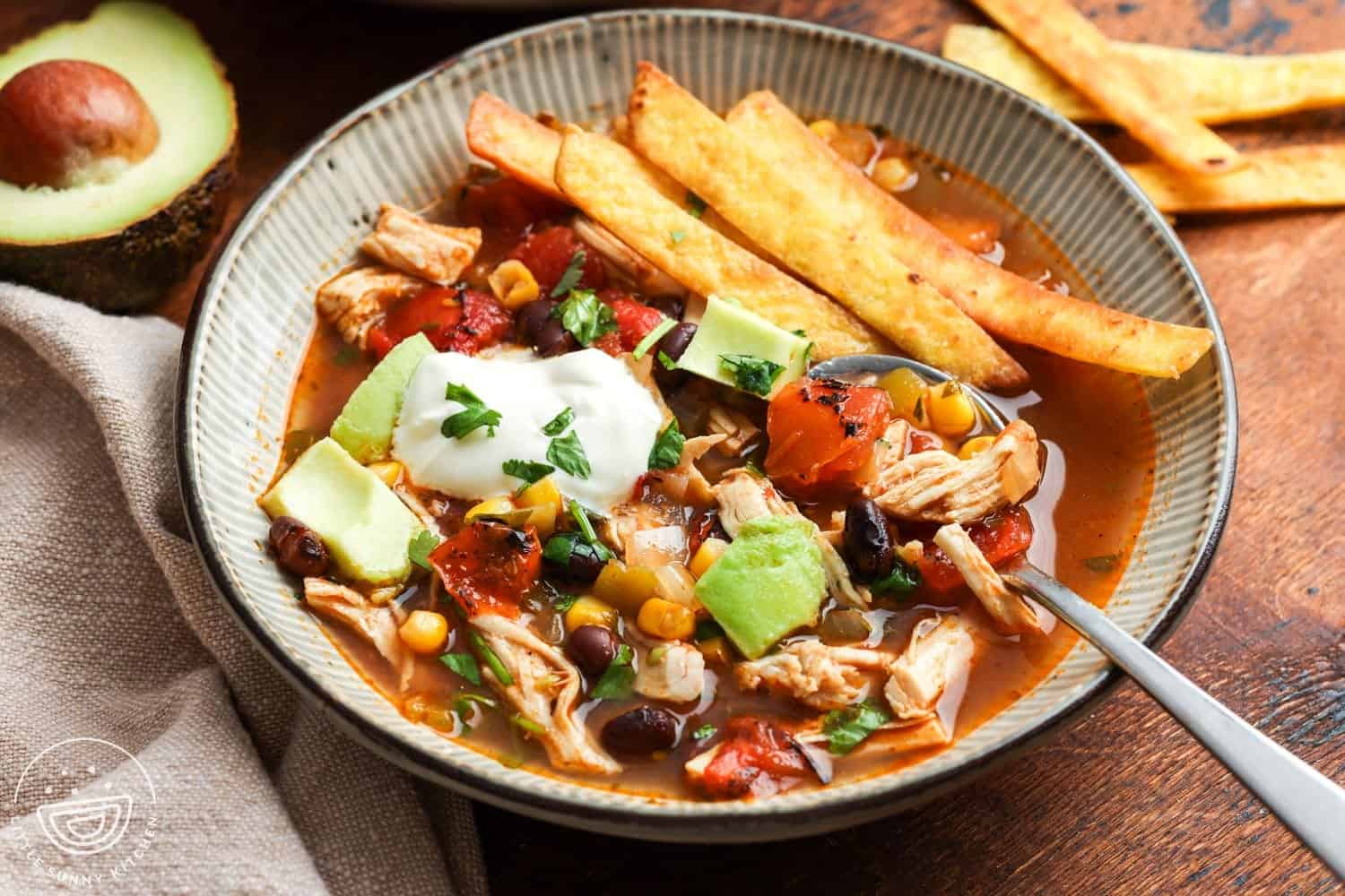 closeup of a bowl of chicken tortilla soup topped with sour cream and diced avocado, on a wooden table. In the background is half an avocado and extra fried tortilla strips.