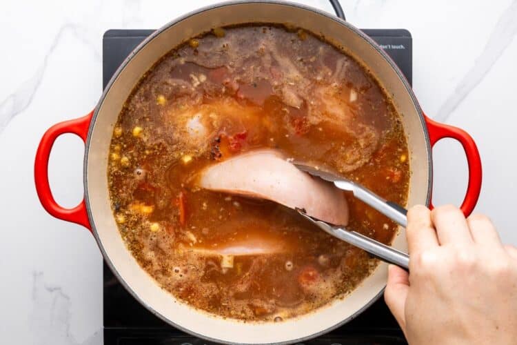 raw chicken breasts added to a pot of soup with tongs.