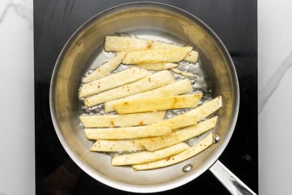 sliced corn tortillas frying in oil in a stainless steel skillet.