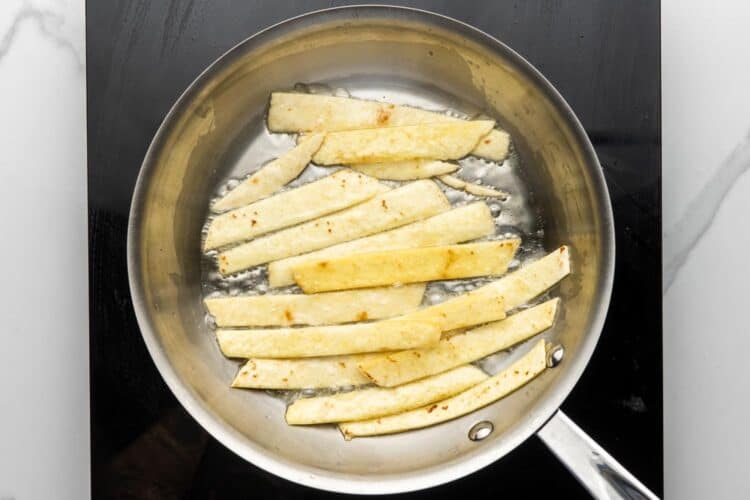 sliced corn tortillas frying in oil in a stainless steel skillet.
