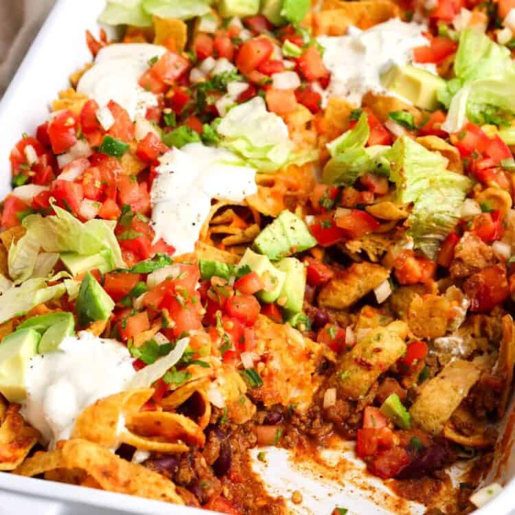 closeup of a frito pie casserole with layers of beef, beans, chips, cheese, and fresh toppings, including sour cream and avocado.