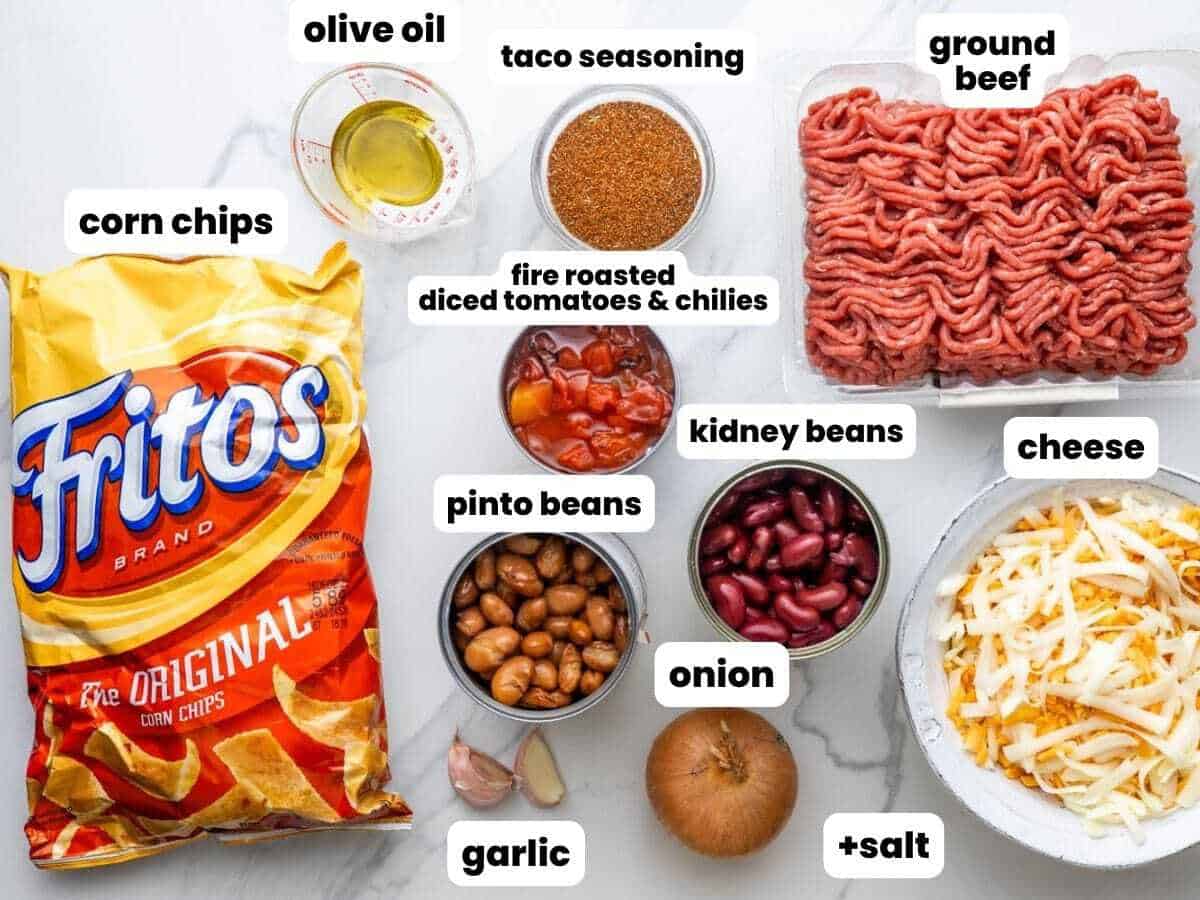 Ingredients for making frito pie casserole, including canned beans and ground beef, arranged on a marble countertop