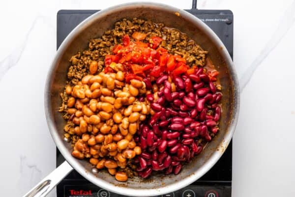 two types of beans and canned tomatoes added to seasoned ground beef in a skillet.