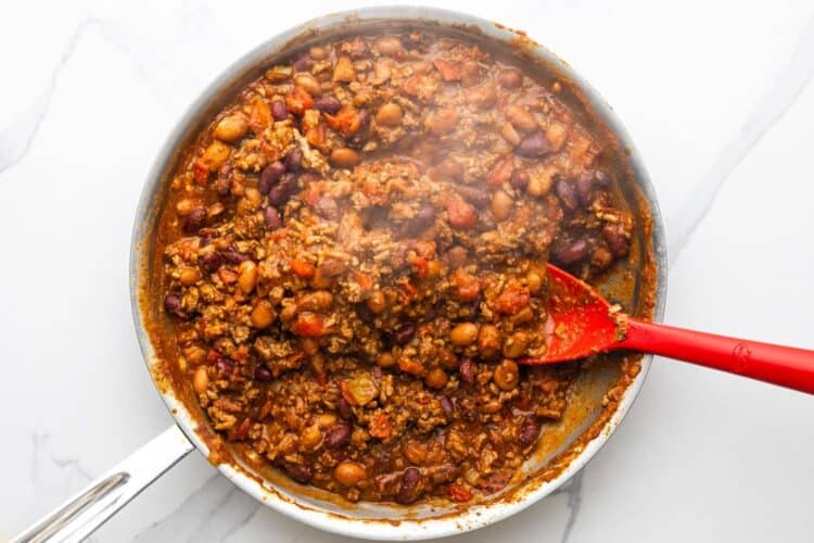 beef and bean chili for frito pie, simmering in a skillet, stirred with a red spoon.