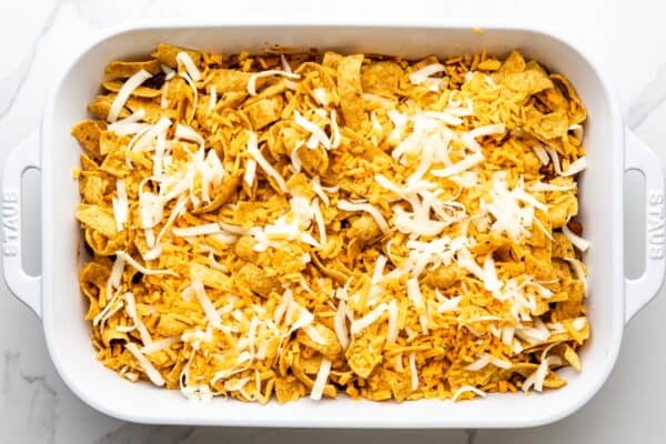 crushed Frito chips and shredded cheese added to the top of a frito pie casserole in a white ceramic dish.