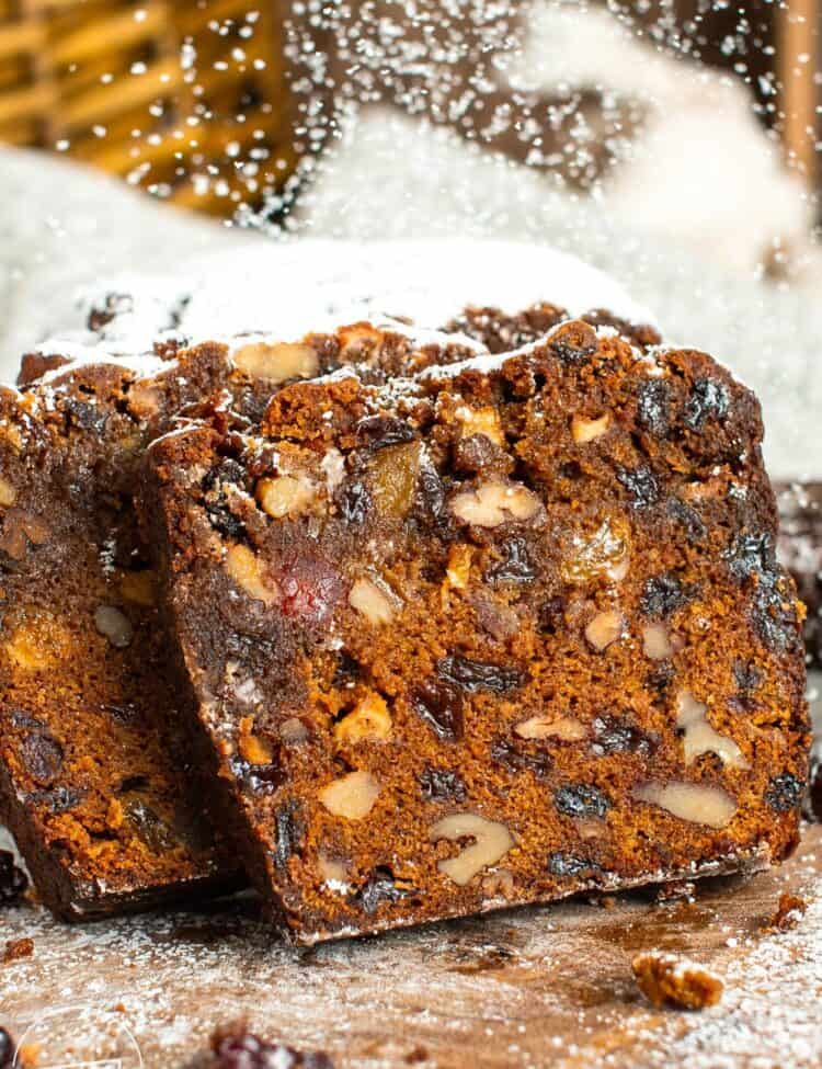 Two slices of fruit cake leaned against the loaf. Inside each slice is plenty of dried fruit and walnuts. Powdered sugar is dusting over the bread.