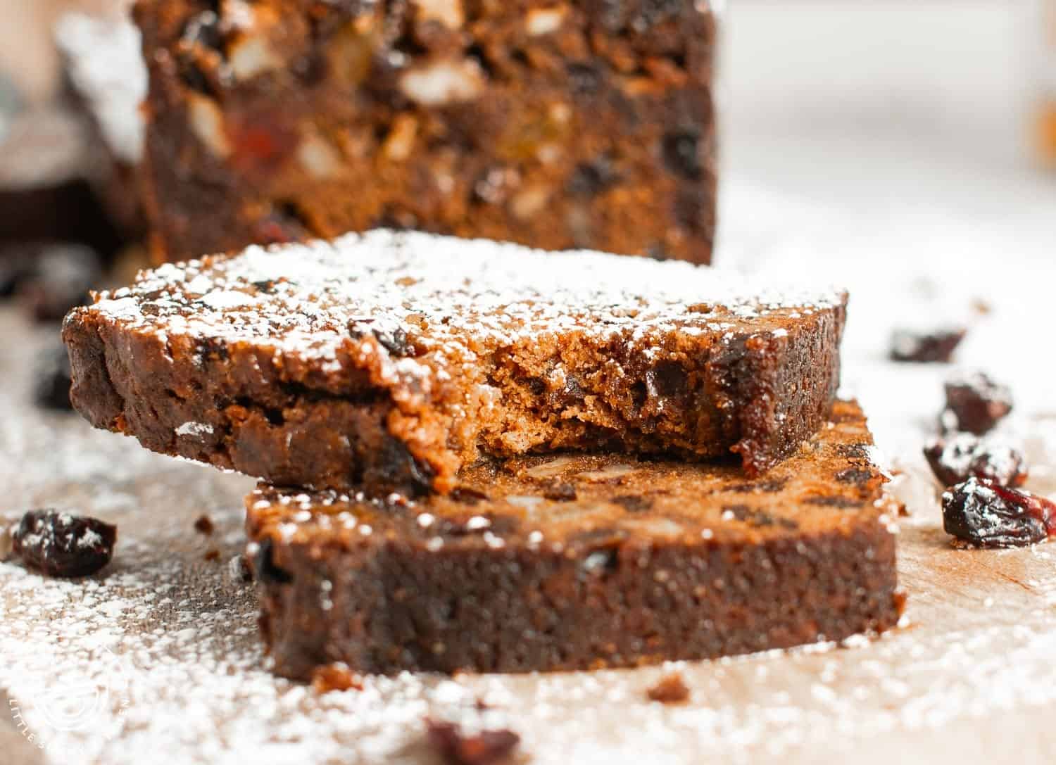 Two slices of fruitcake, stacked. The top slice is dusted with powdered sugar and has a bite taken out of it to show the tender texture.