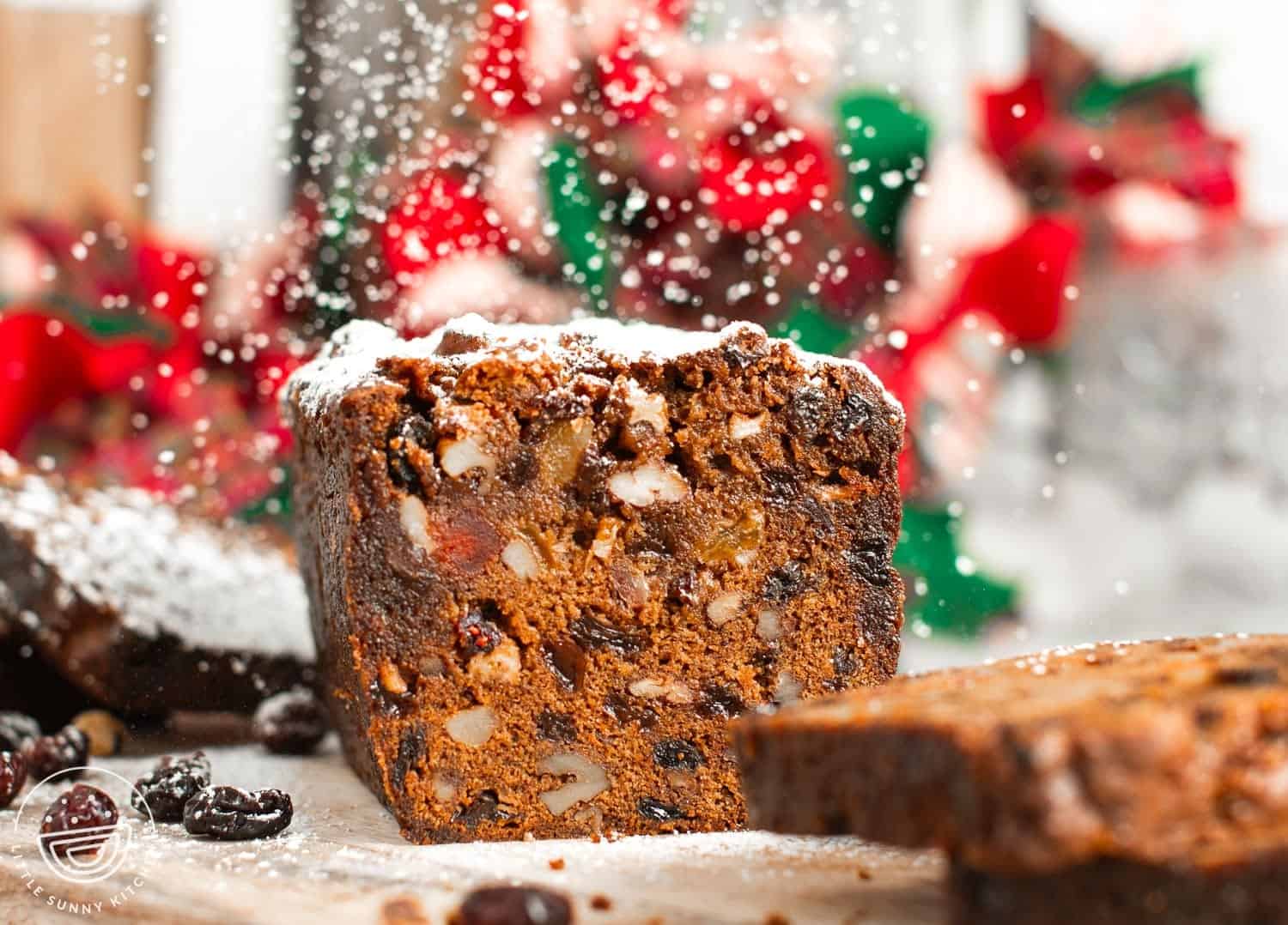 a loaf of fruitcake, sliced in half with powdered sugar dusted over it like snow. blurred, in the background are red and green holiday decorations.
