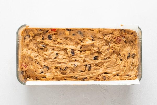 thick fruitcake batter is spread into a parchment lined loaf pan.