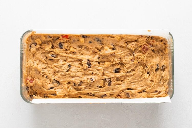 thick fruitcake batter is spread into a parchment lined loaf pan.