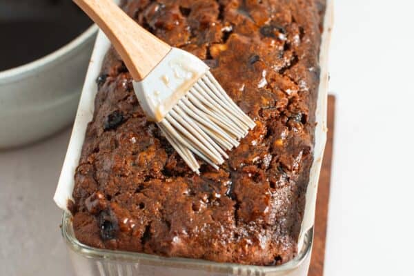 a silicone pastry brush adding syrup to the top of a baked fruit cake in a glass pan.