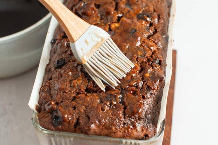 a silicone pastry brush adding syrup to the top of a baked fruit cake in a glass pan.