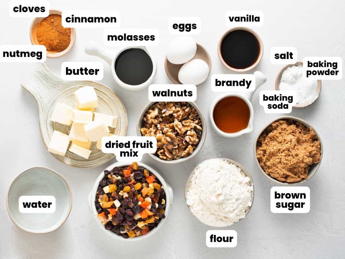 The ingredients needed to make a classic fruit cake with dried fruits, walnuts, molasses and brandy. All of the cake ingredients are measured into small bowls, arranged on white surface.