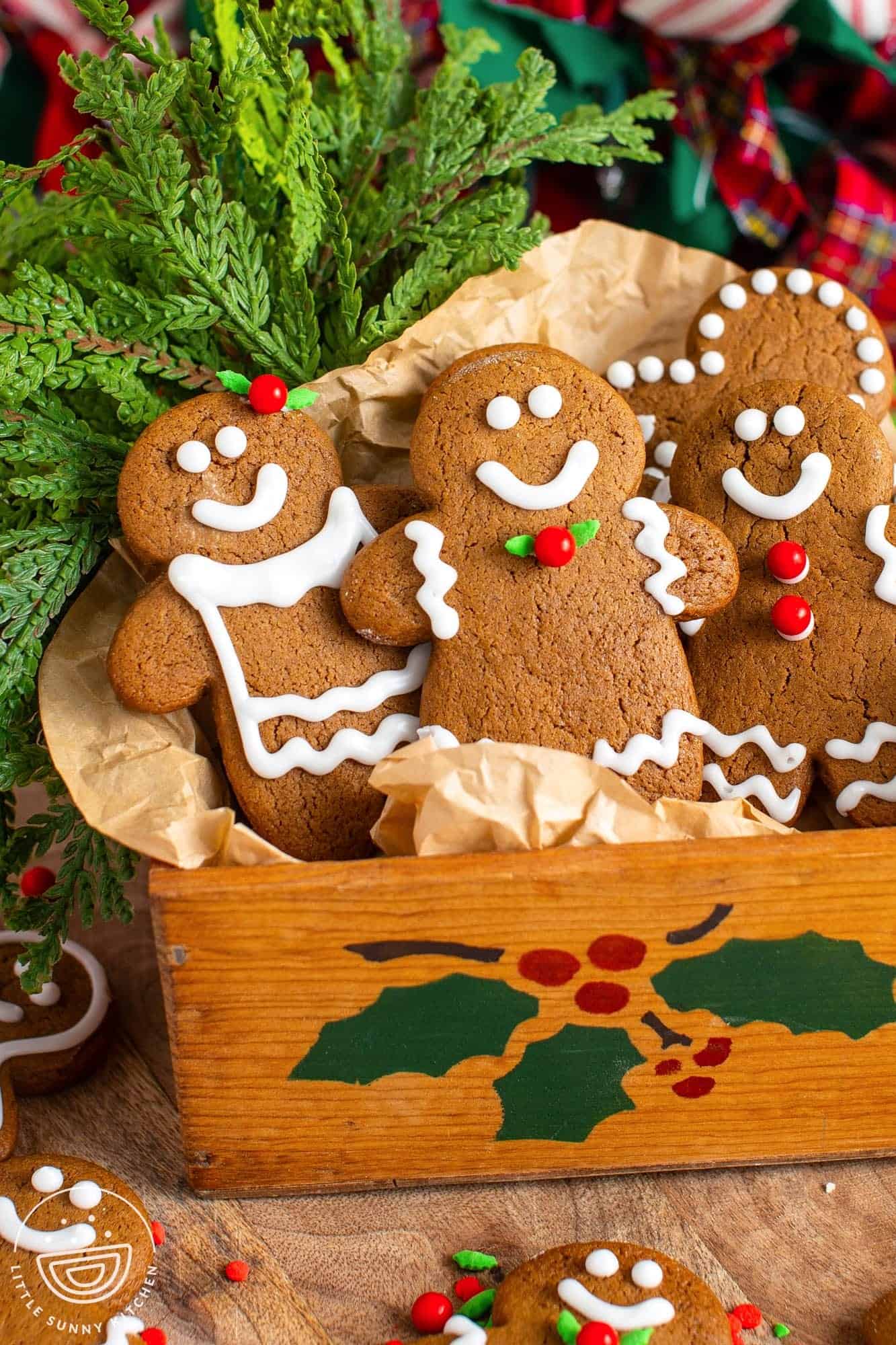 decorated gingerbread men arranged in a holiday painted wooden box. In the background is greenery and red plaid ribbon.