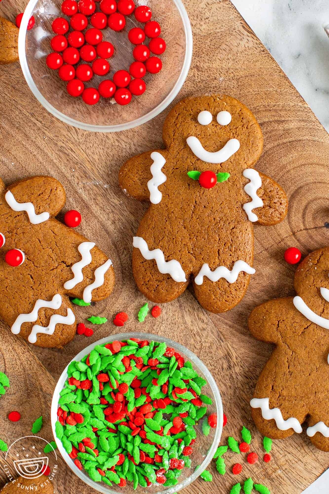 large gingerbread man cookies arranged on a wooden board along with small clear bowls of holiday sprinkles. The cookies are decorated in the traditional way with white icing.