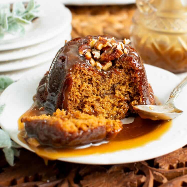 small sticky toffee pudding on a white plate with toffee sauce and chopped pecans.