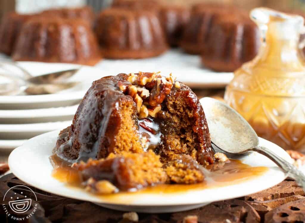 an individual sized sticky toffee pudding on a small white plate with a spoon. The cake has been split open to show the nuts and sticky caramel inside.