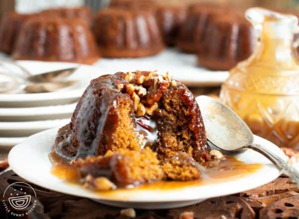 an individual sized sticky toffee pudding on a small white plate with a spoon. The cake has been split open to show the nuts and sticky caramel inside.