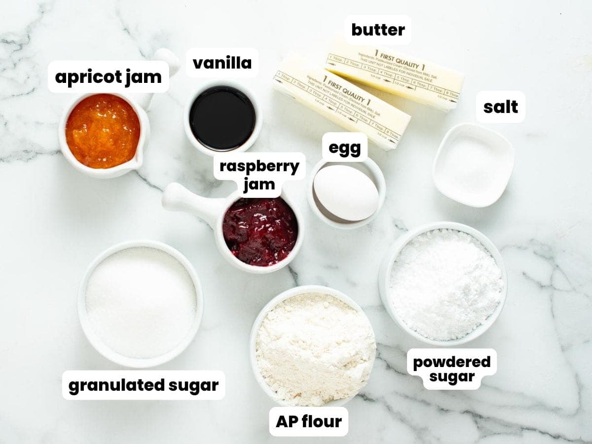 Ingredients for making thumbprint cookies with apricot and raspberry jam, two sticks of butter, flour, sugar, egg, vanilla, and salt. Each ingredient is measured into a small white bowl and set on a marble counter.