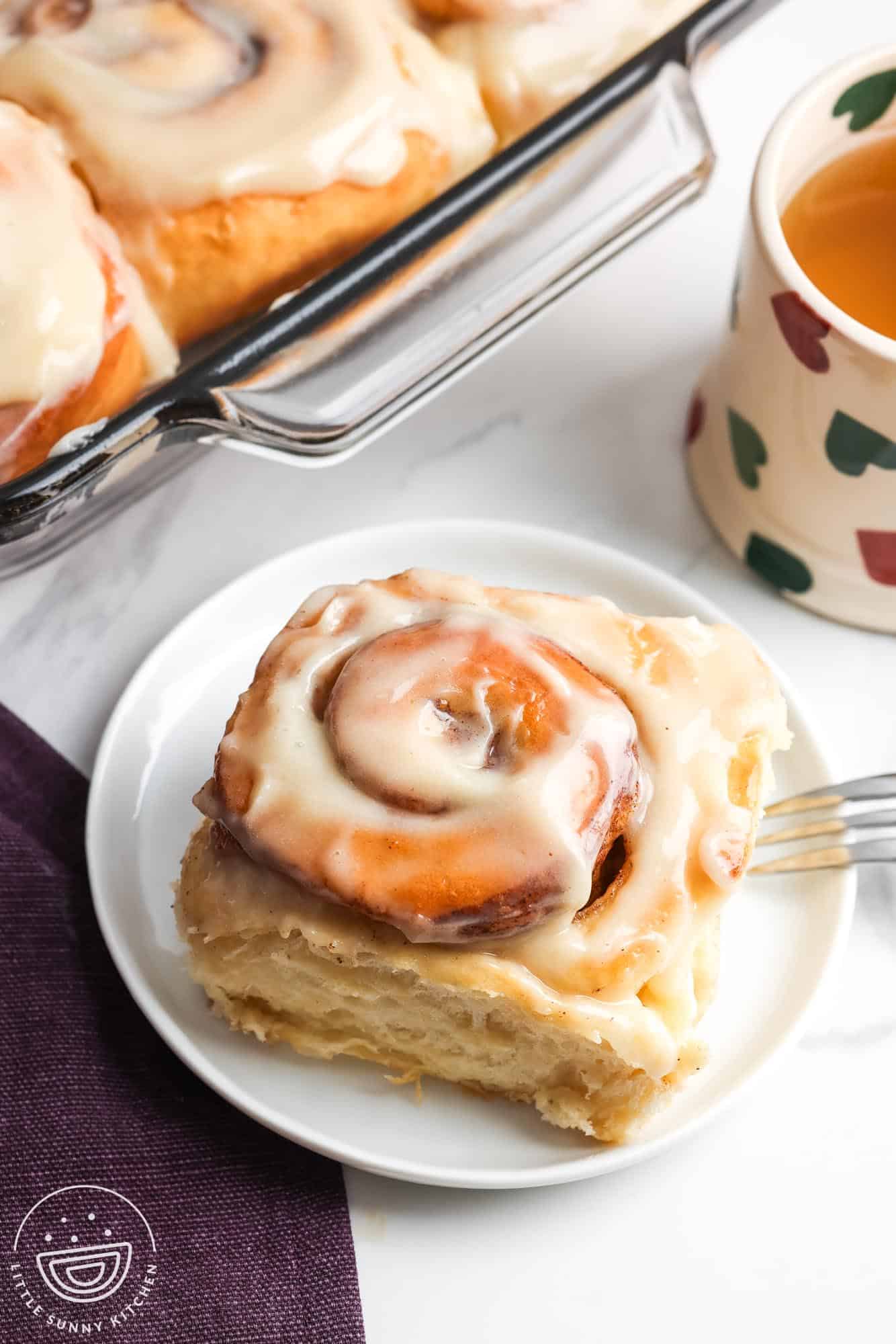 A cinnamon roll with icing on a small white plate next to a mug of tea and a pan of cinnamon rolls.