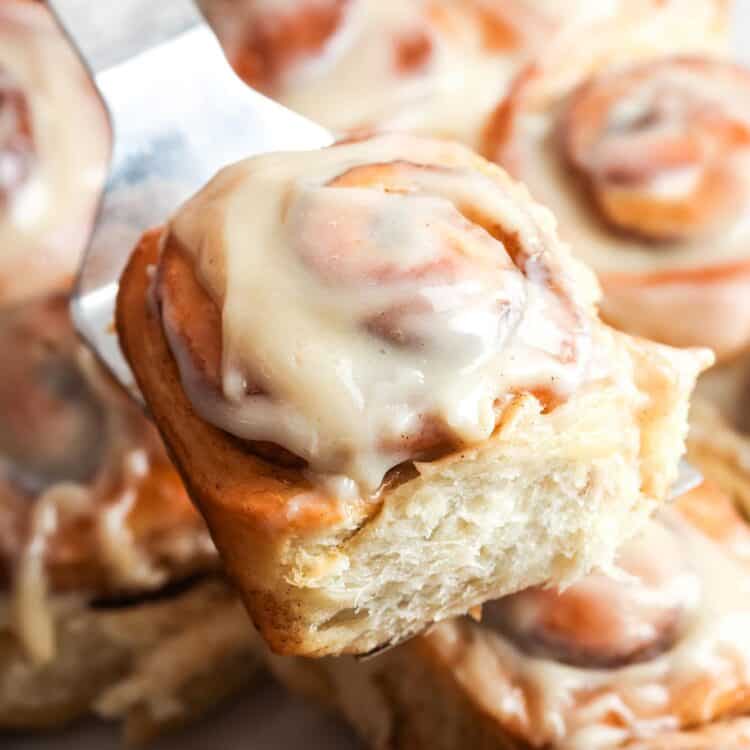 a fluffy frosted cinnamon roll lifted from a pan of rolls with a metal spatula.