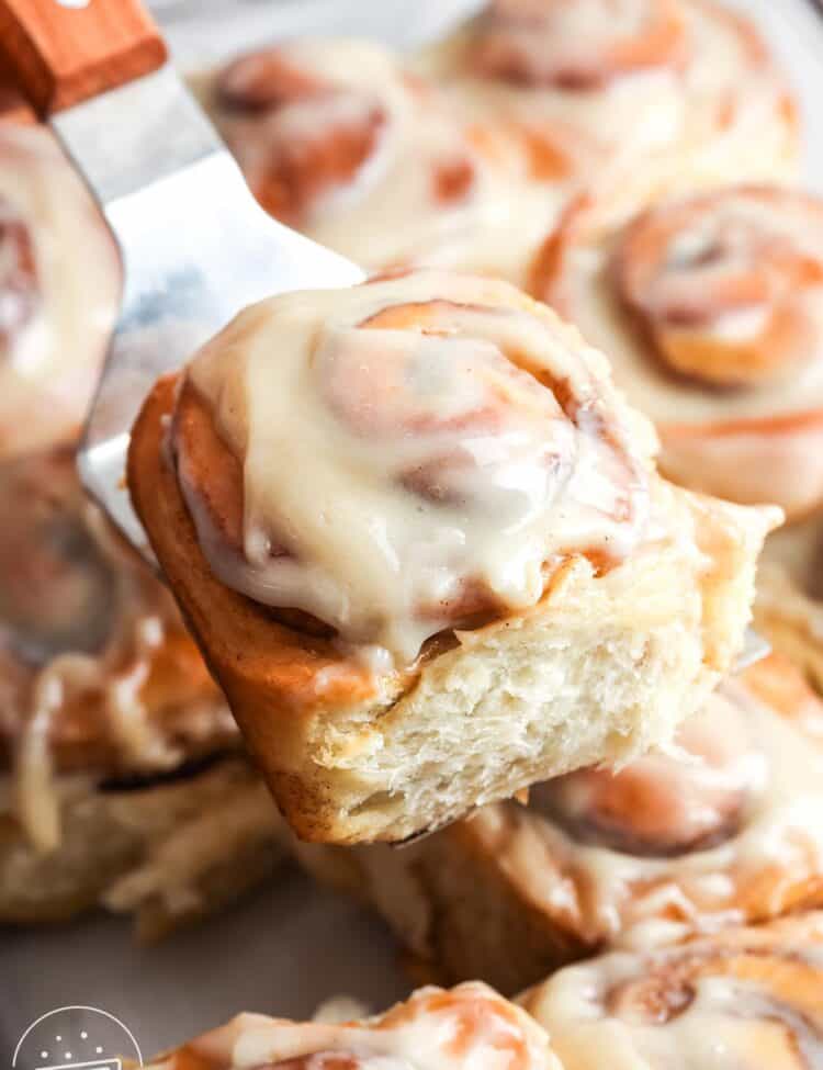 a fluffy frosted cinnamon roll lifted from a pan of rolls with a metal spatula.