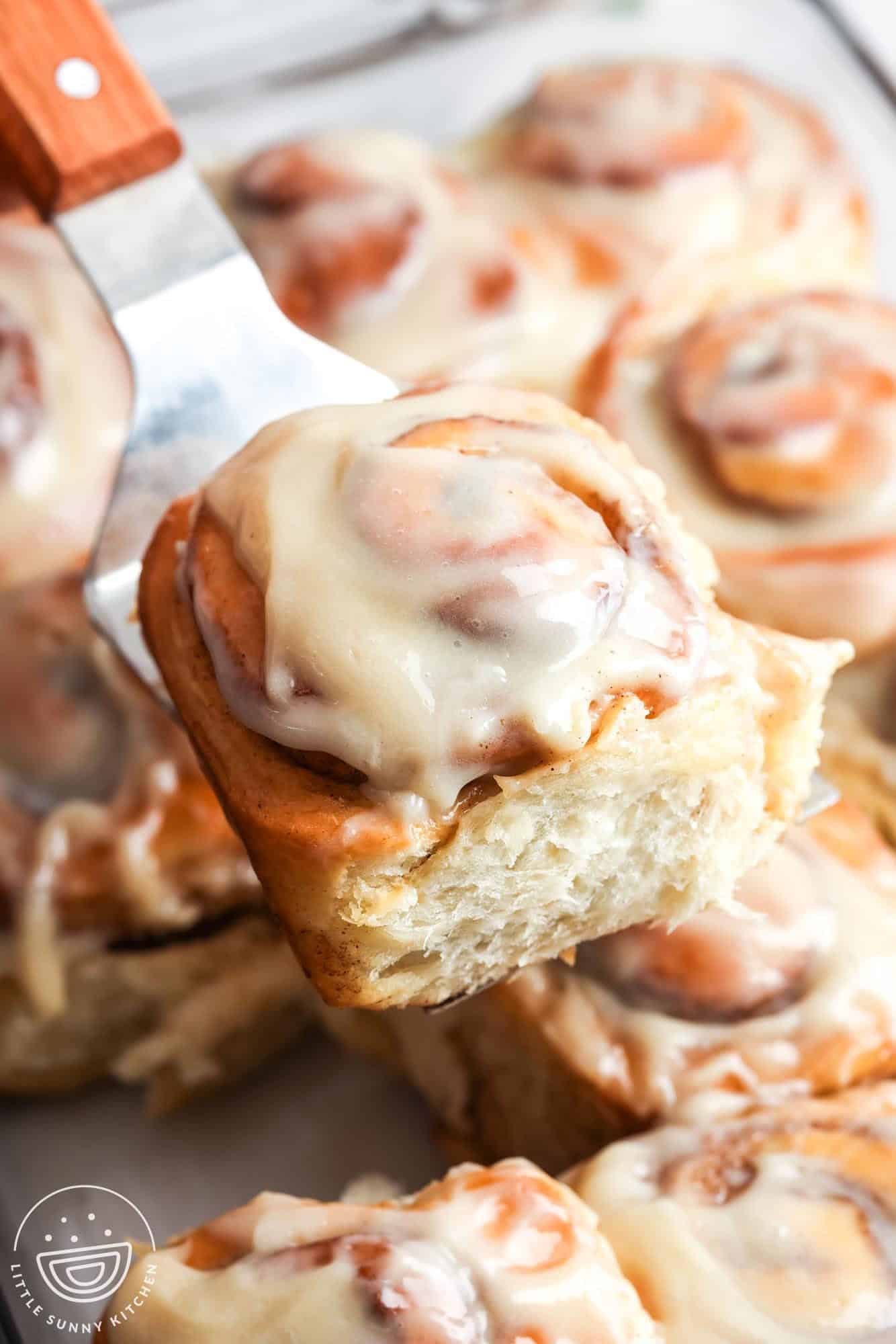 a fluffy frosted cinnamon roll lifted from a pan of rolls with a metal spatula.