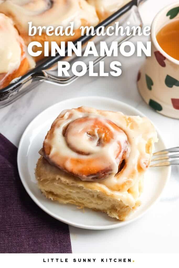 an iced cinnamon roll on a small white plate next to a mug of tea. Text overlay says "bread machine cinnamon rolls"