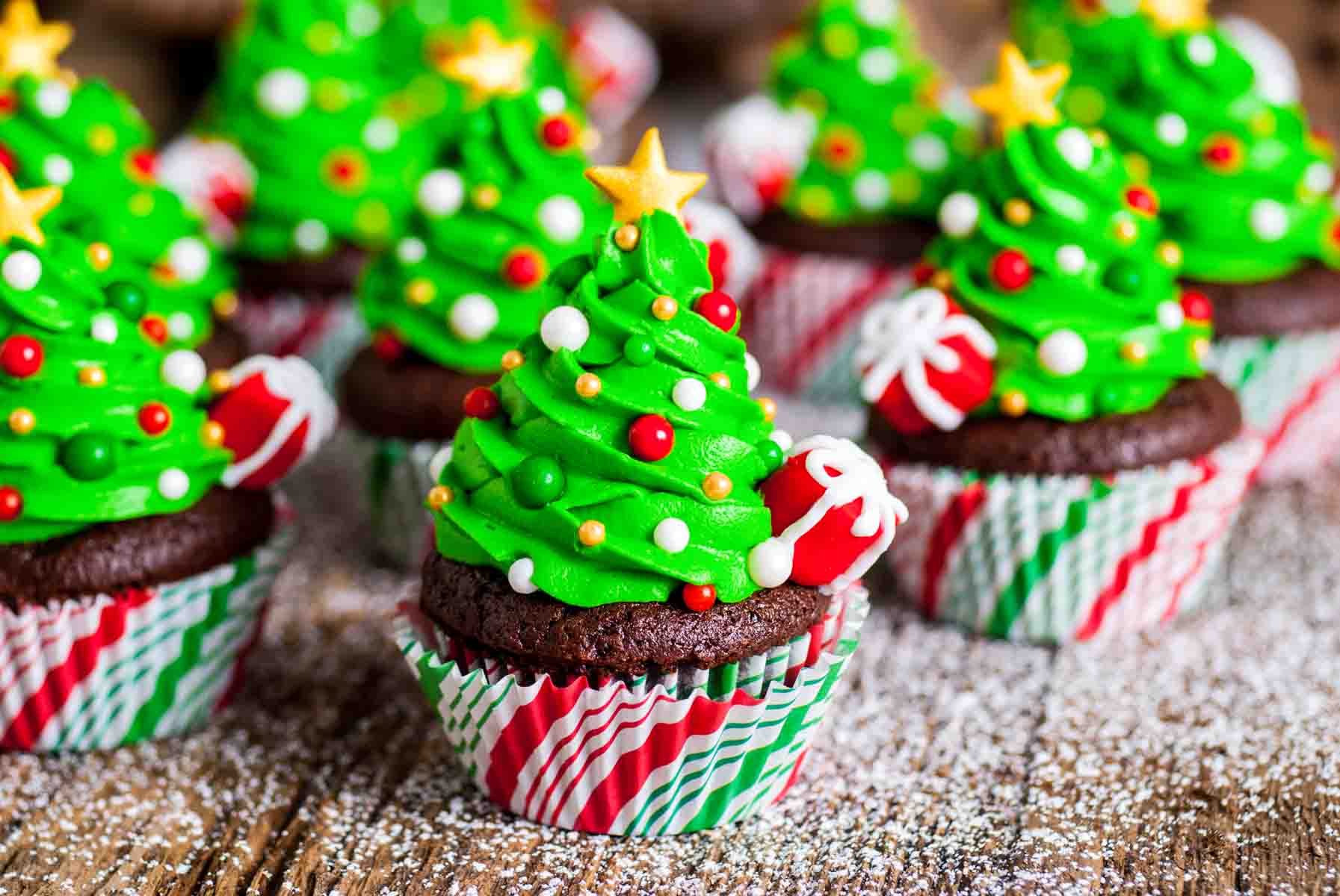 Chocolate cupcakes topped with bright green Christmas tree frosting decorated with sprinkles and a star topper on a wooden surface dusted with powdered sugar