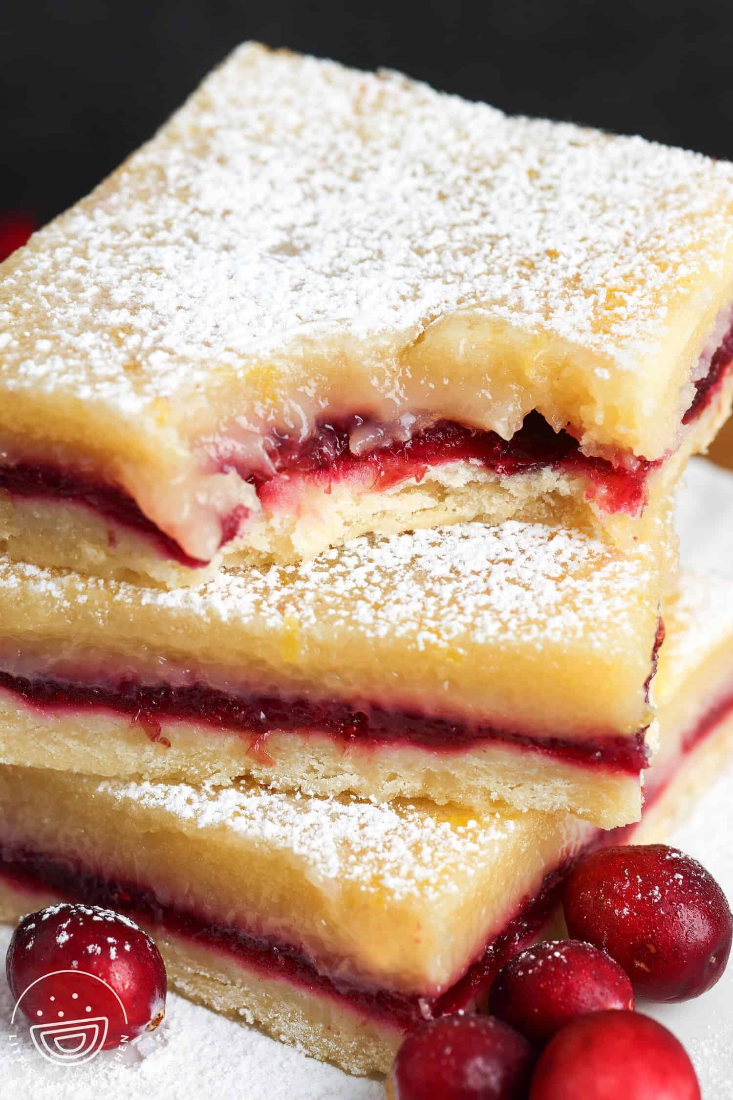 three cranberry lemon bars stacked. They are topped with powdered sugar. One bar has a bite taken to show the soft lemon curd and flaky crust.