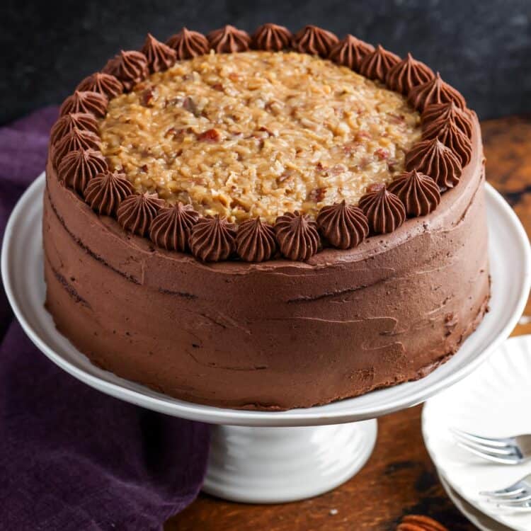 A german chocolate layer cake topped with coconut, on a white cake stand set on a wooden table next to a dark purple towel.
