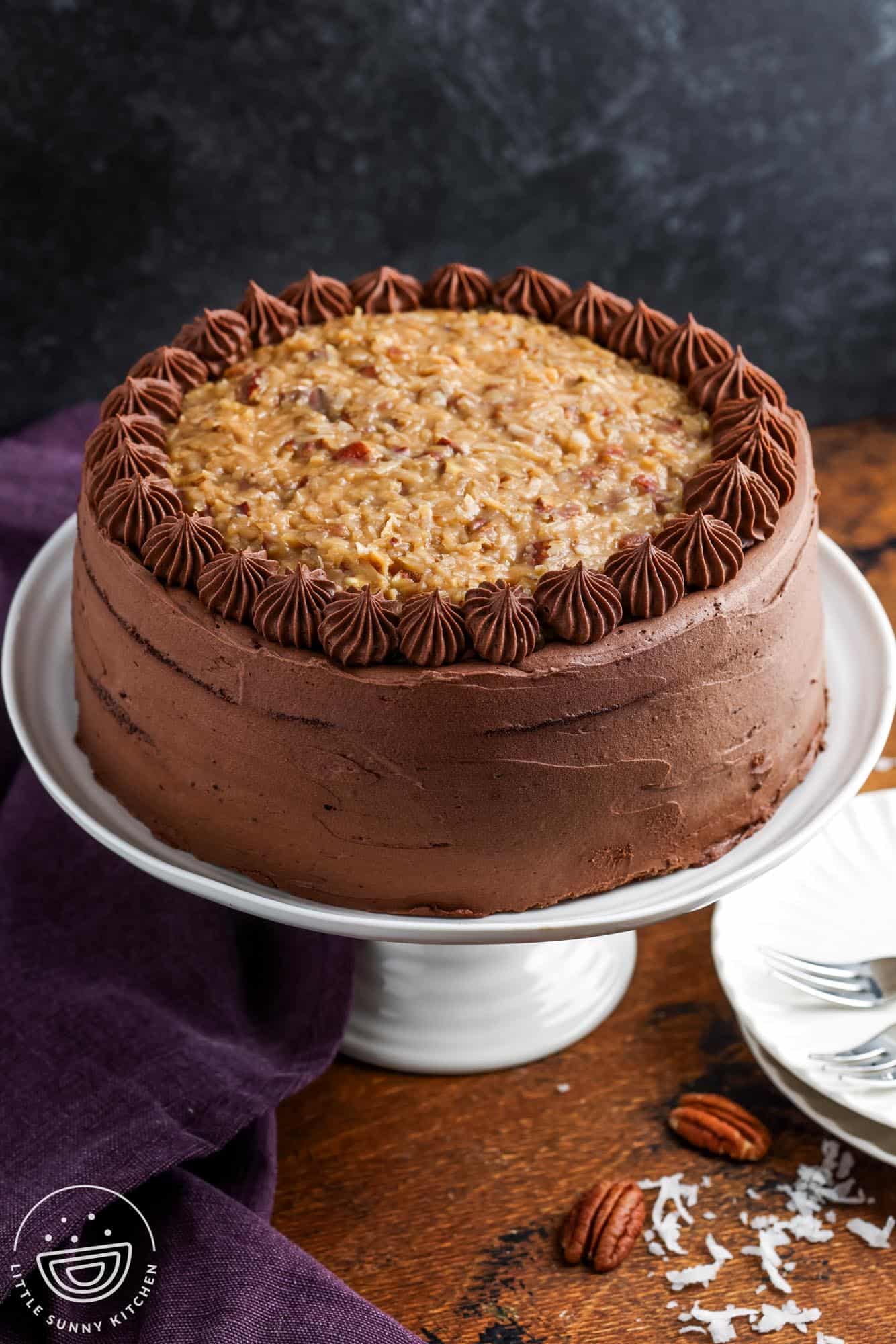 A german chocolate layer cake topped with coconut, on a white cake stand set on a wooden table next to a dark purple towel.