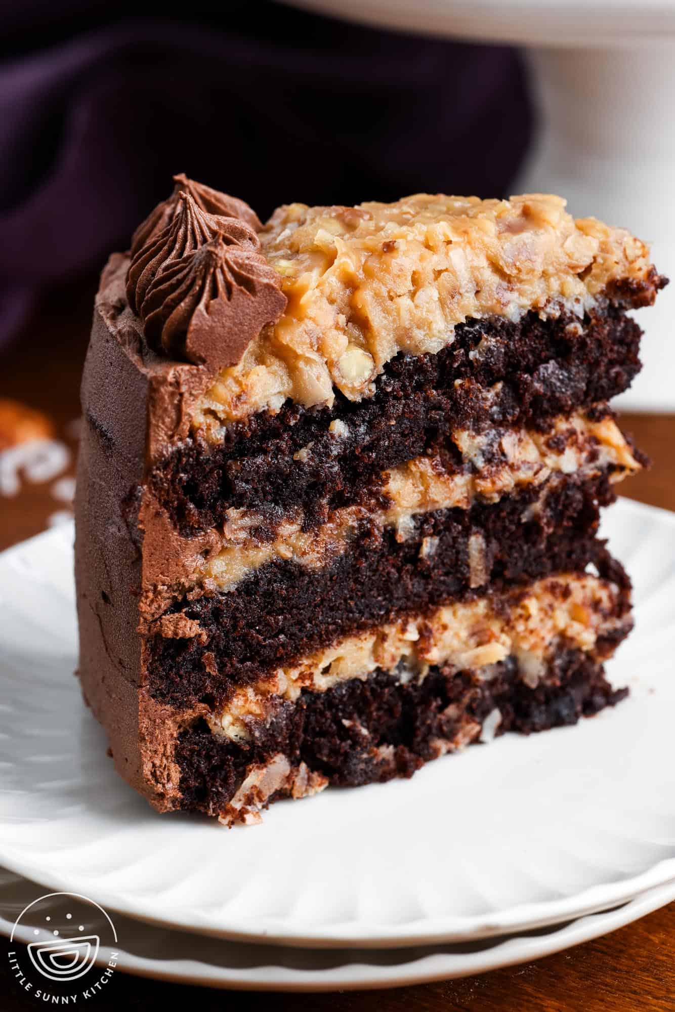 one slice of german chocolate cake set on a small white plate. The cake slice is three layers, filled with creamy coconut pecan filling and iced with chocolate buttercream.
